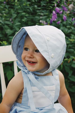 baby in blue and white bonnet with white buttons in front of green leafy background