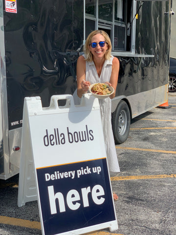 Julie is standing in front of the mobile kitchen, holding a bowl, over the sign that reads "della bowls: pick up deliveries here."