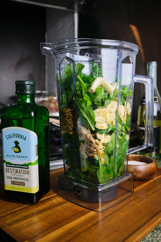 A blender full of ingredients for green sauce sits on a wooden cutting board. 