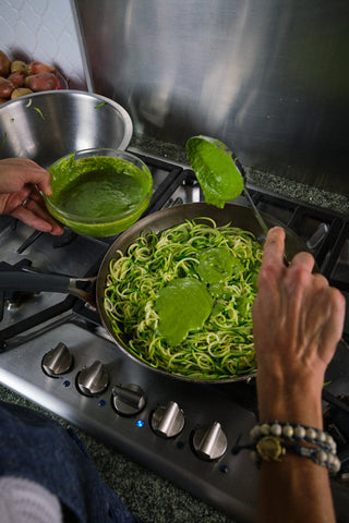 zucchini noodles are cooking in a skillet and a hand is adding green sauce as they cook