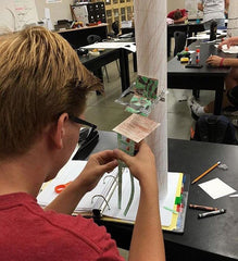 A student constructs a model deer stand at his desk using paper, colored pencils, and tape.