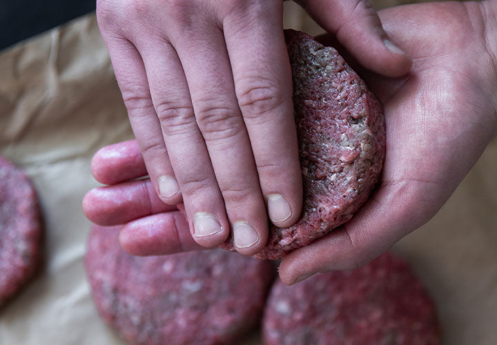 Hands forming ground beef into a hamburger patty next to finished hamburger patties.