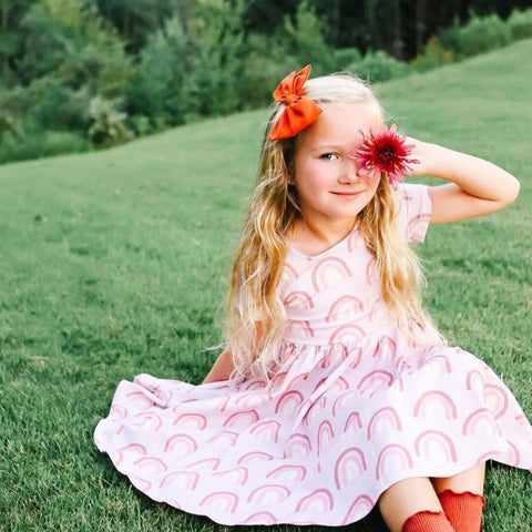 girl sitting on grass holding flower over eye in rainbow twirl dress