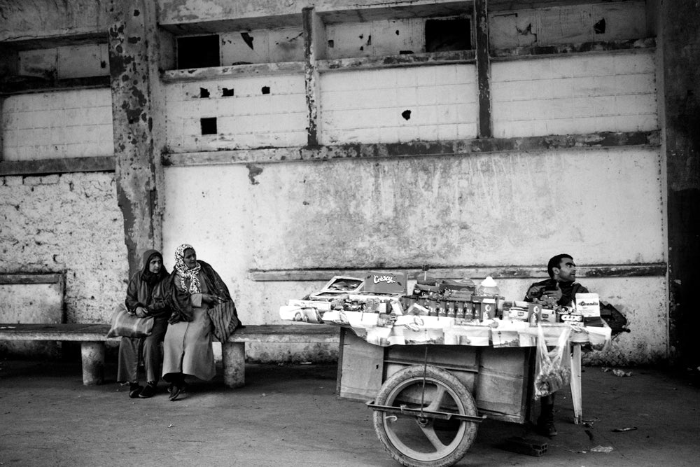 Two women on cement bench wait for bus next to a street vendor. Fez, Morocco.