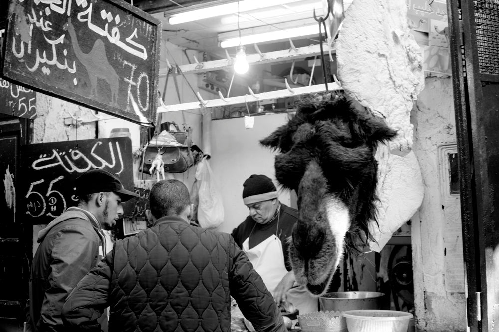 A butcher standing beside a hanging camel head serves two customers inside his shop.