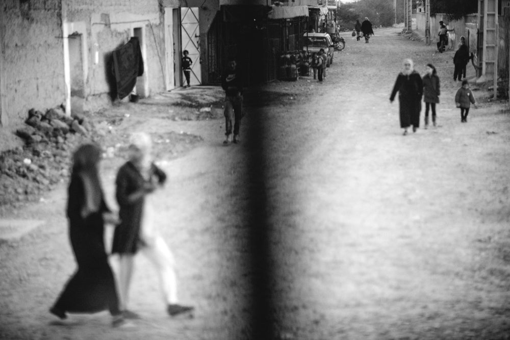 Villagers and townspeople walk in a wide street.  Image captured through a bus window while in transit to Chefchaouen.