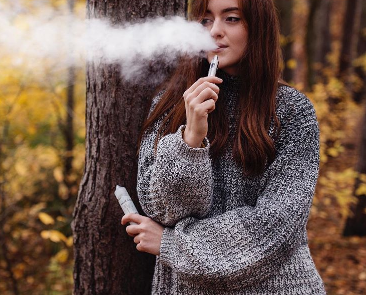 a woman leans against the tree as she blows smoke from her mouth while vaping