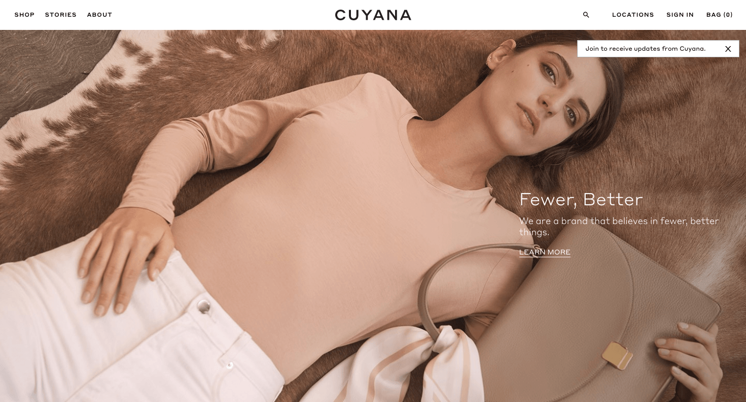 cuyana's fewer better campaign hero image of woman lying on a cowhide rug wearing a blush pink shirt and white jeans and holding a beige tote bag
