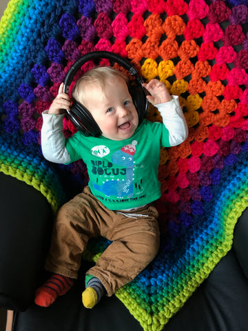 Elliott sits on a black chair, wearing head phones, on a chunky rainbow crochet blanket
