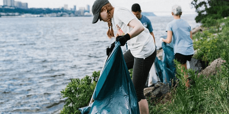 A woman holding a trash bag and cleaning litter from the shore line.