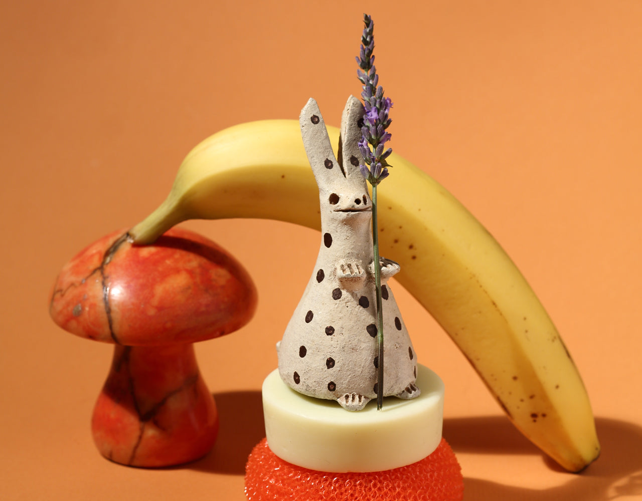 Unwrapped conditioner bar in a stack of a round red scrub sponge and a rabbit sculpture with brown polka-dots holding a stalk of lavender. There's a red mushroom sculpture and banana behind the rabbit, and a peach background.
