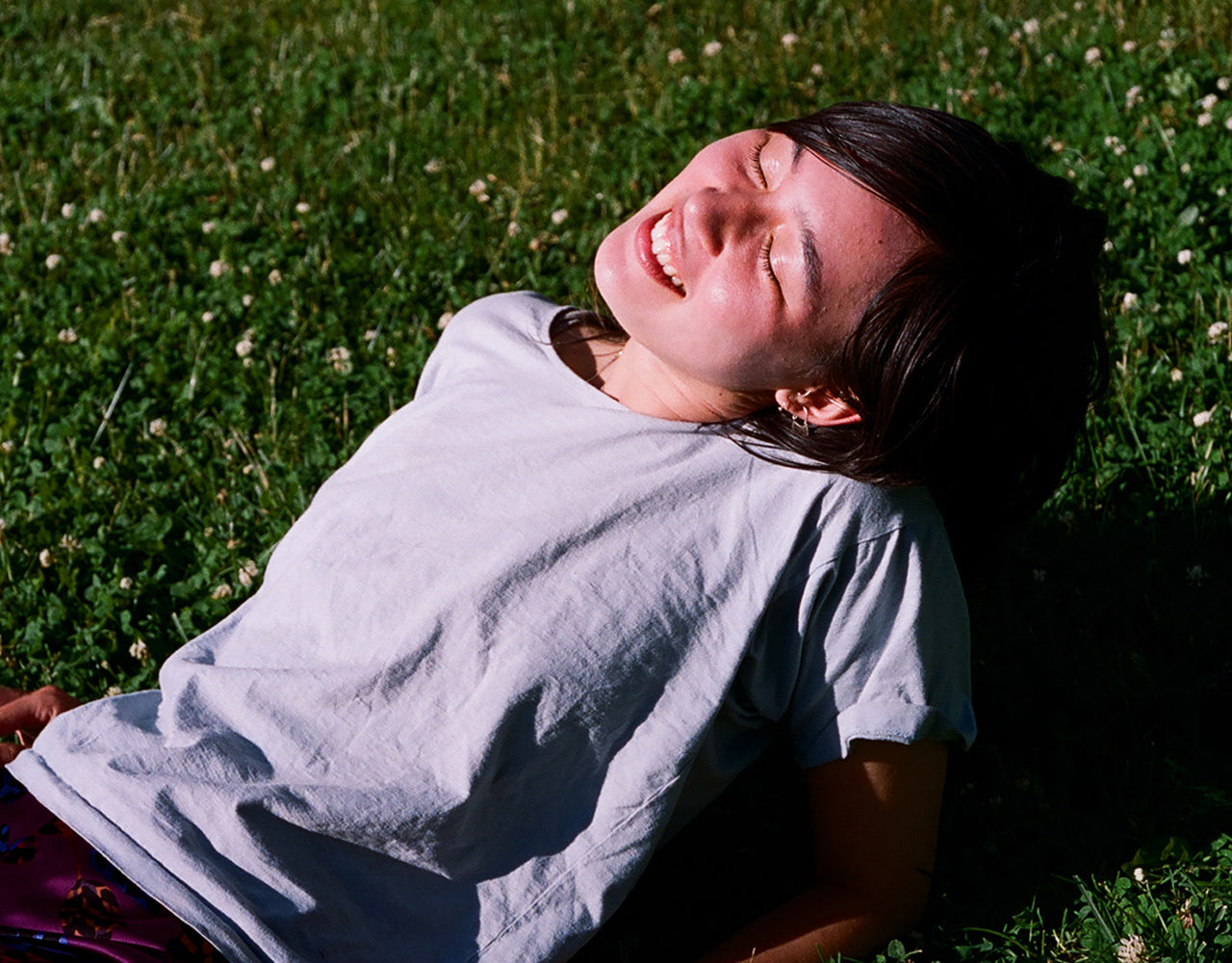 Person with black hair and a white t-shirt lying on the their back on the grass in full sun