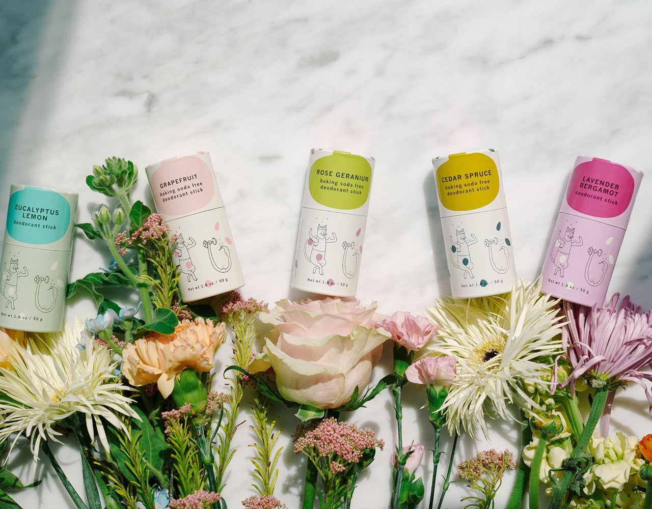 Meow Meow Tweet deodorant sticks laying on a white marble background with several wildflowers below.