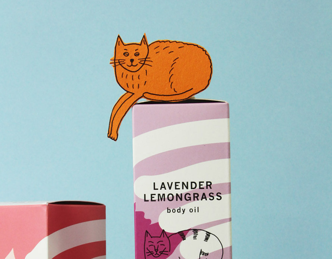 hand-drawn orange paper cat cutout on top of a box of Meow Meow Tweet Lavender Lemongrass body oil against a light blue background