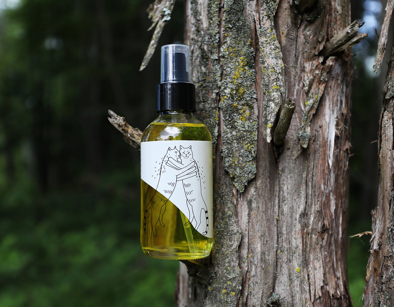 A bottle of Lemon Rose Cleansing oil screenprinted with two cats standing on hind legs grooming each other. The bottle is in a tree with shredded bark, moss, and lichen.