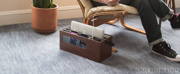 Walnut wood charging station resting on carpet in living room next to designer recliner where man uses phone while it charges