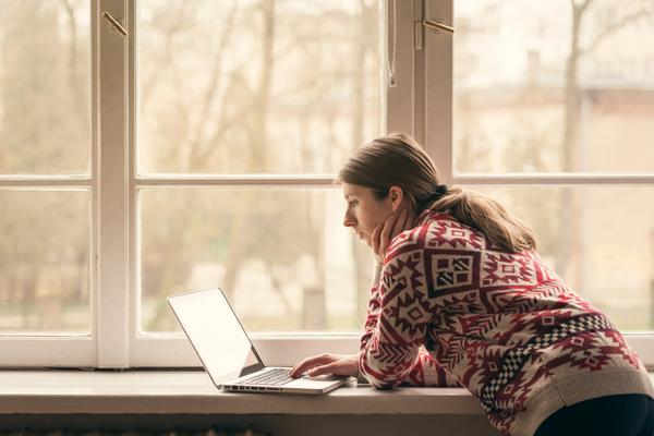 A-woman-in-a-knit-sweater-stands-using-her-MacBook-on-the-window-sill-in-winter