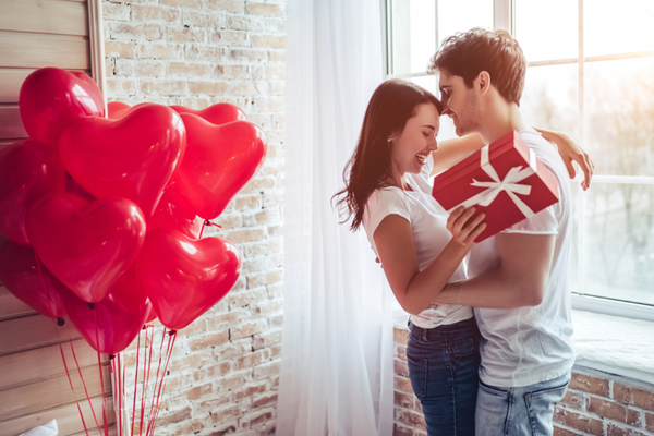 A-young-couple-embraces,-enjoying-Valentine’s-Day-The-woman-is-holding-a-gift-box-and-they-are-standing-next-to-heart-shaped-balloons.