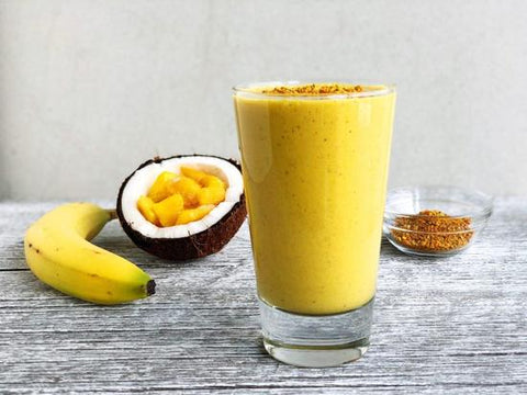 a tropical turmeric smoothie topped with bee pollen in front of a banana, a halved coconut filled with mango and a glass of bee pollen