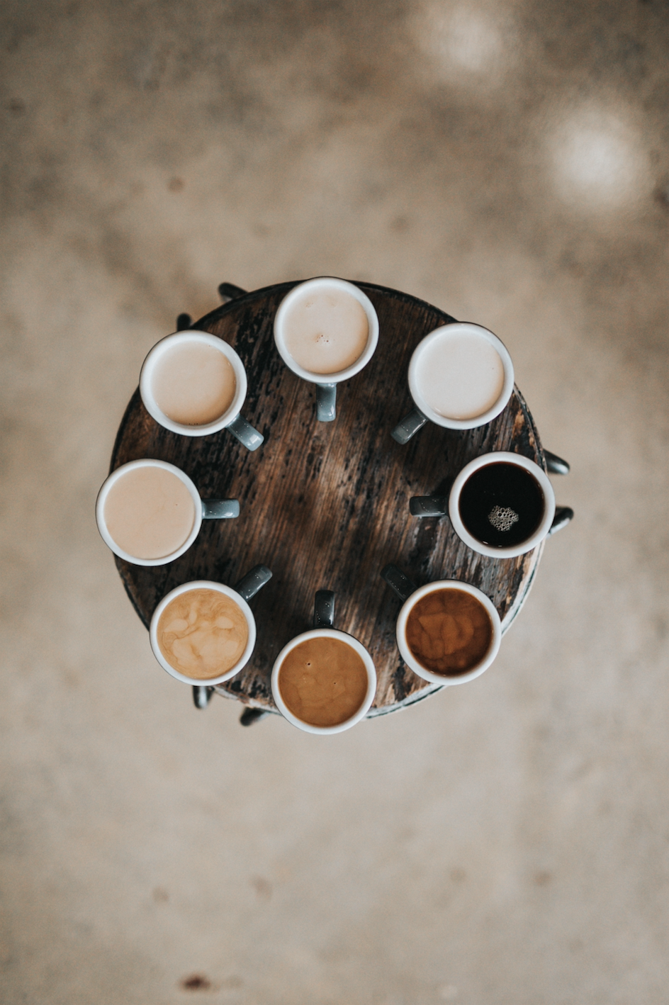 Aerial view of eight coffee cups on a round table filled with coffee and each with a different amount of milk added to create varying coffee colors. 