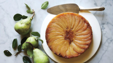 pear upside down cake on a white platter with a silver serving lifter next to ripe green pears with leaves