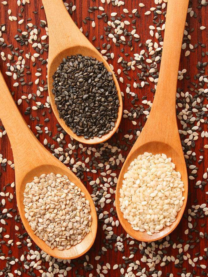 Three wooden spoons filled with black and white sesame seeds lie on a red wooden backdrop surrounded by sprinkled sesame seeds. 