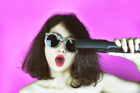 woman with damaged hair wearing sunglasses straightening her hair with flat iron on purple background
