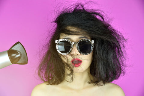 woman wearing sunglasses on purple background blow-drying her hair