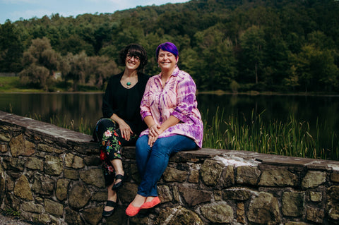 Lauren Beacham and Brittany Elbourn of Yugen Tribe in outdoor setting by a lake sitting on a stone wall
