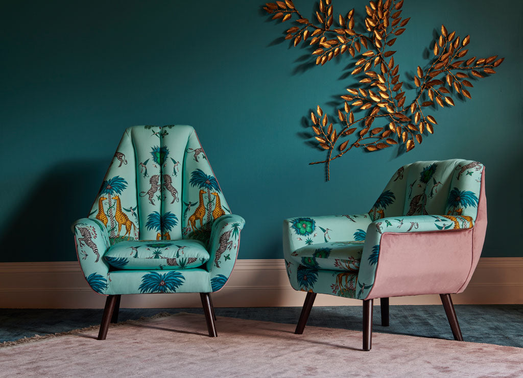 Two chairs upholstered in the Creatura Turquoise Cotton Satin Fabric, from the Wilderie collection by Emma J Shipley with Clarke & Clarke. Featuring leopard spotted giraffes and prancing zebras under a mystical palm tree, injecting animal magic into your home interior