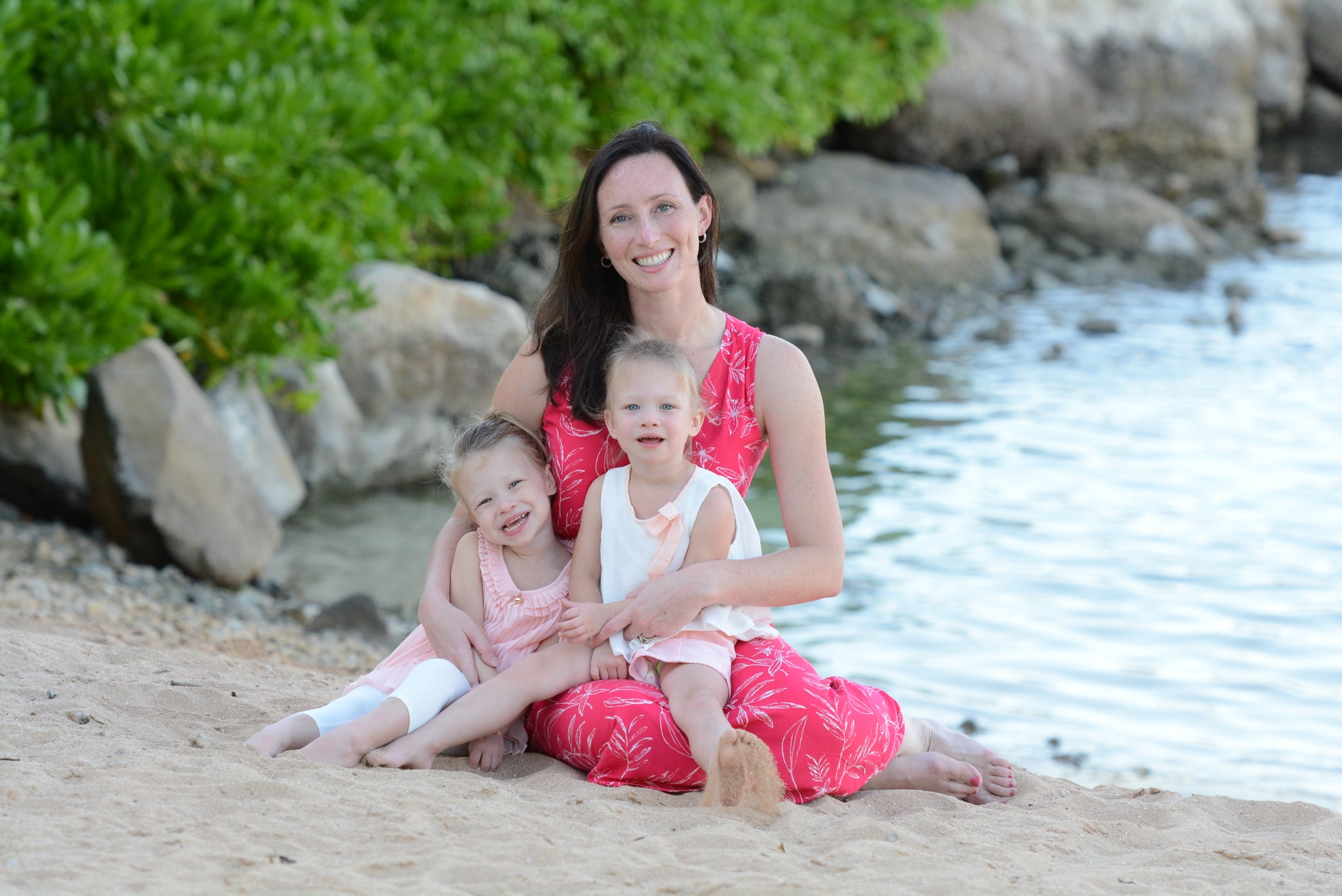 Woman in pink dress with two young girls sitting on a beach.