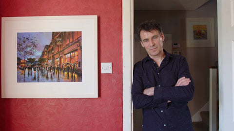 Photo of artist Chris McMorrow standing beside a print of one of his paintings of Clerys Clock, Dublin