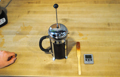 Press pot sitting while it brews the coffee, next to a spoon and a timer