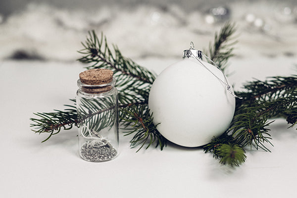A white Christmas bauble on a white surface with green twigs and a pot of glitter