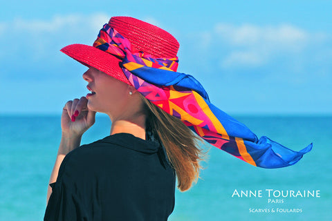 An ANNE TOURAINE Paris™ multicolor silk scarf tied around a red straw hat 
