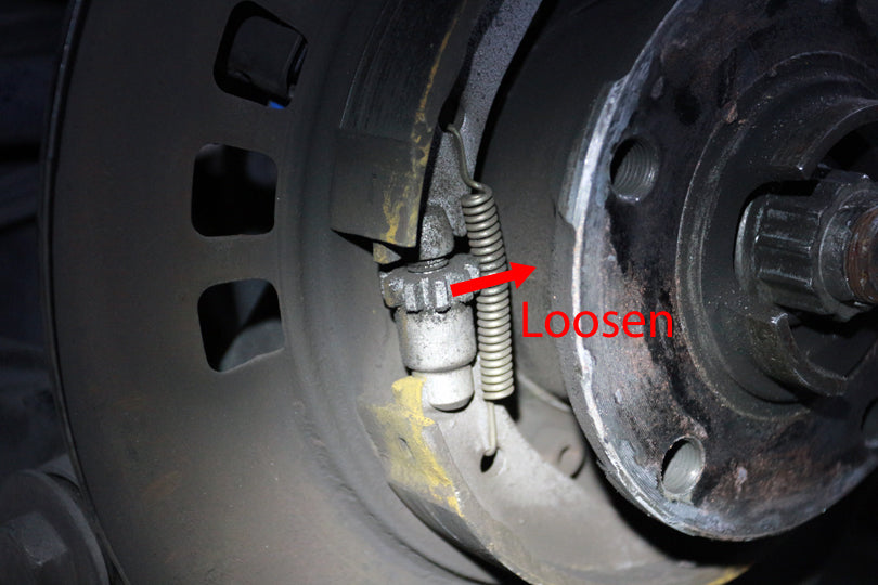 Parking brake adjustment mechanism with arrow showing that rotating the adjustment mechanism towards the hub loosens the mechanism.