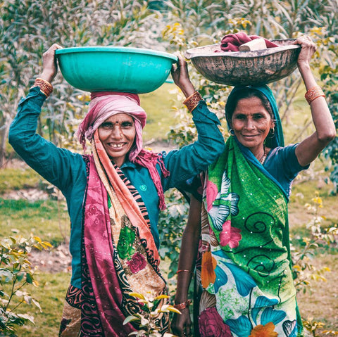Two Indian women dressed in vibrant clothing, carrying baskets of fabric on their heads.