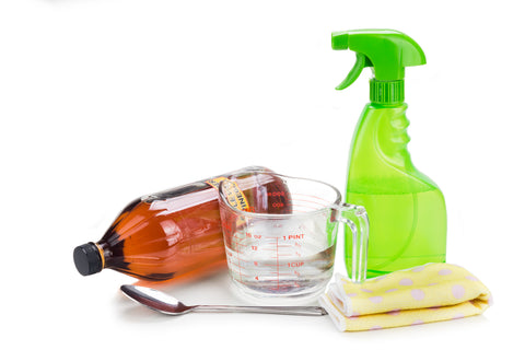 A spray bottle, measuring cup, cleaning rag, vinegar container and spoon stand in front of a white background