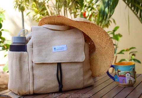 Backpack is set on a wood table with a straw hat on top of it. A colorful coffee mug is next to it and some flowers are in the background.
