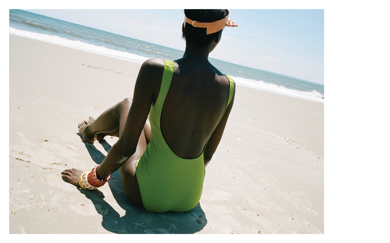 Image of a woman sitting on the beach wearing a Green one pice Swimsuit by Nu Swim with bangles and a visor by Hatori.