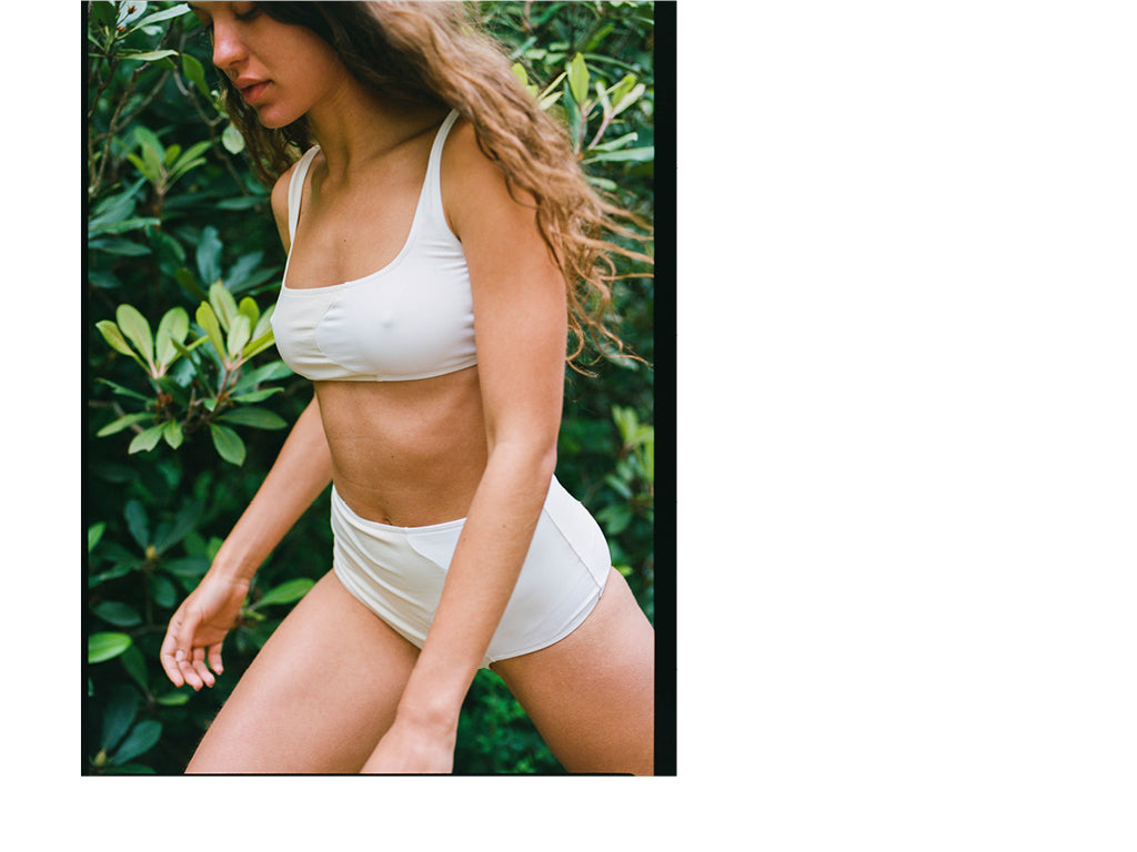 A woman is walking past green bushes wearing a Cream and White bikini by Laura Urbinati.