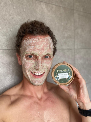 A smiling man with exfoliating face mask on holding Deerieo Secret Garden natural face mask tub.