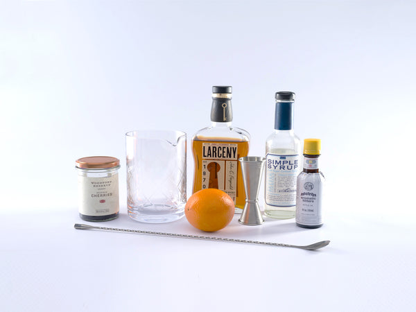 Ingredients for an old fashioned: From left to right - Jar of bourbon glazed cherries, mixing glass, Larceny Bourbon, Simple syrup, bitters, large metal spoon and an orange.  