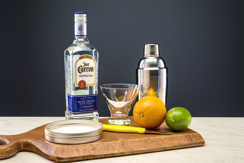 Ingredients for a skinny margarita on a wooden cutting board. From Left to right: Coarse salt, José Cuervo Tequila, margarita glass, paring knife, orange and lime. 