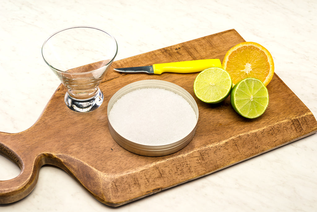 Halved lime and orange on wood cutting board with coarse salt and margarita glass. 