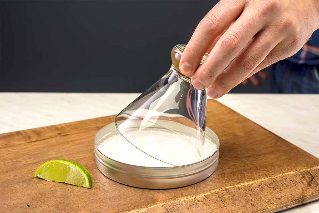 Margarita glass being dipped into tray of coarse salt to salt the rim. 