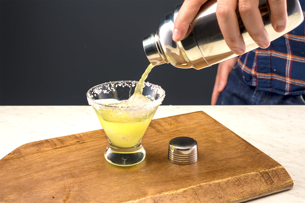 Contents of cocktail shaker (tequila, agave nectar, lime and orange juice) being poured into margarita glass. 