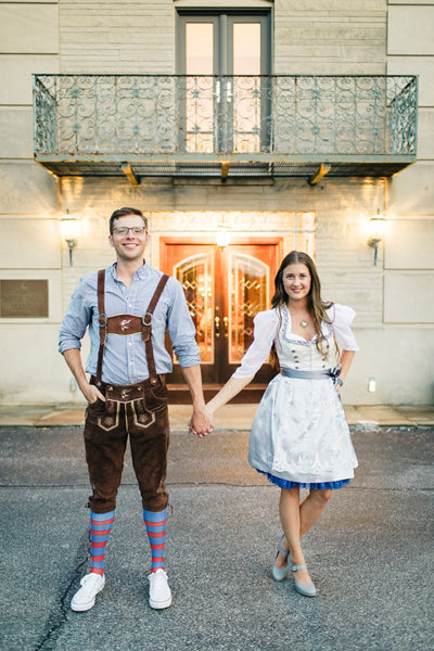 Groom wearing lederhosen standing in front of a fountain with his bride wearing a white and blue wedding dirndl dress for their Oktoberfest rehearsal dinner