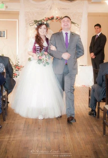 Bride wearing a plum wedding dirndl with a floor length tulle overskirt walking down the aisle with her groom wearing a grey suit