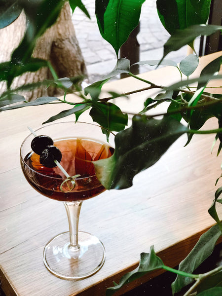 Green leaves and champagne glass on wooden table with brown liquid and cherries on toothpick | DIY Spring Cocktails by Caia Cummings at Batch